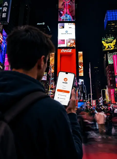 Man holding phone in Times Square, the phone screen shows the Reddit login page
